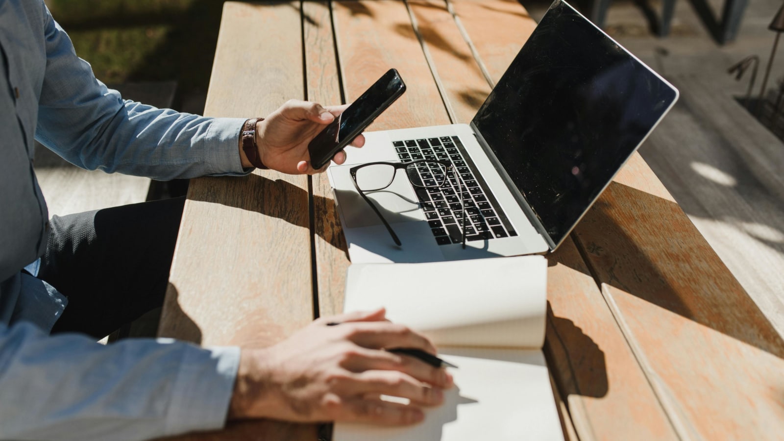 A laptop on a rugged wooden outdoors table and a man working at the table, holding a phone in left hand and writing in a notebook with the right hand.
