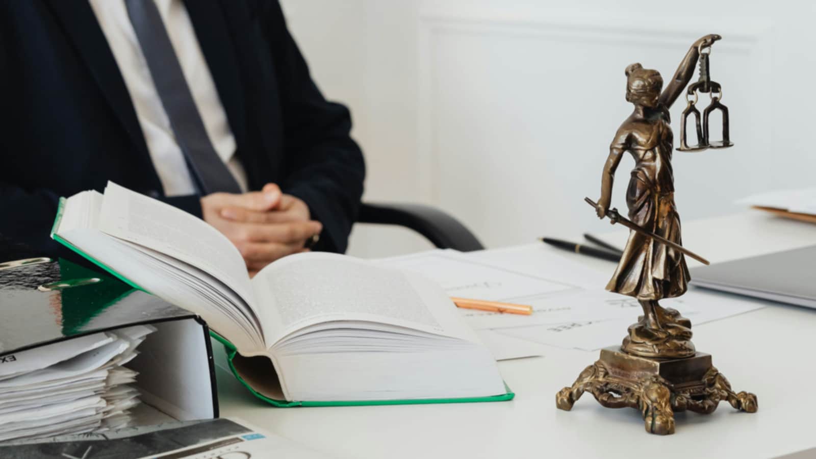 A table with documents, an open law book, and a miniature statue of Lady Justitia on it. A man with a suit and tie sitting behind the desk. 
