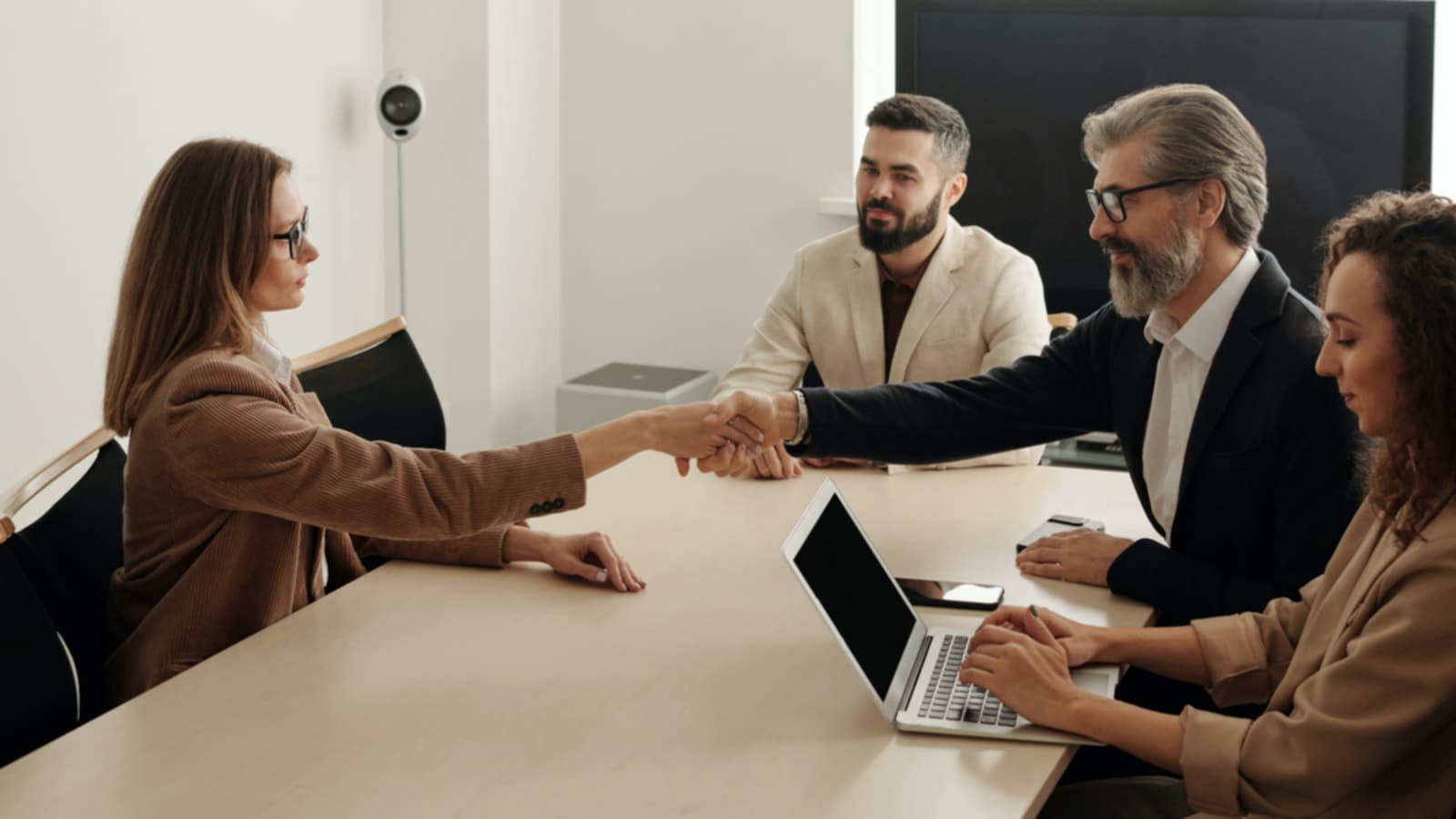 A handshake over a meeting table, four people in a meeting. 