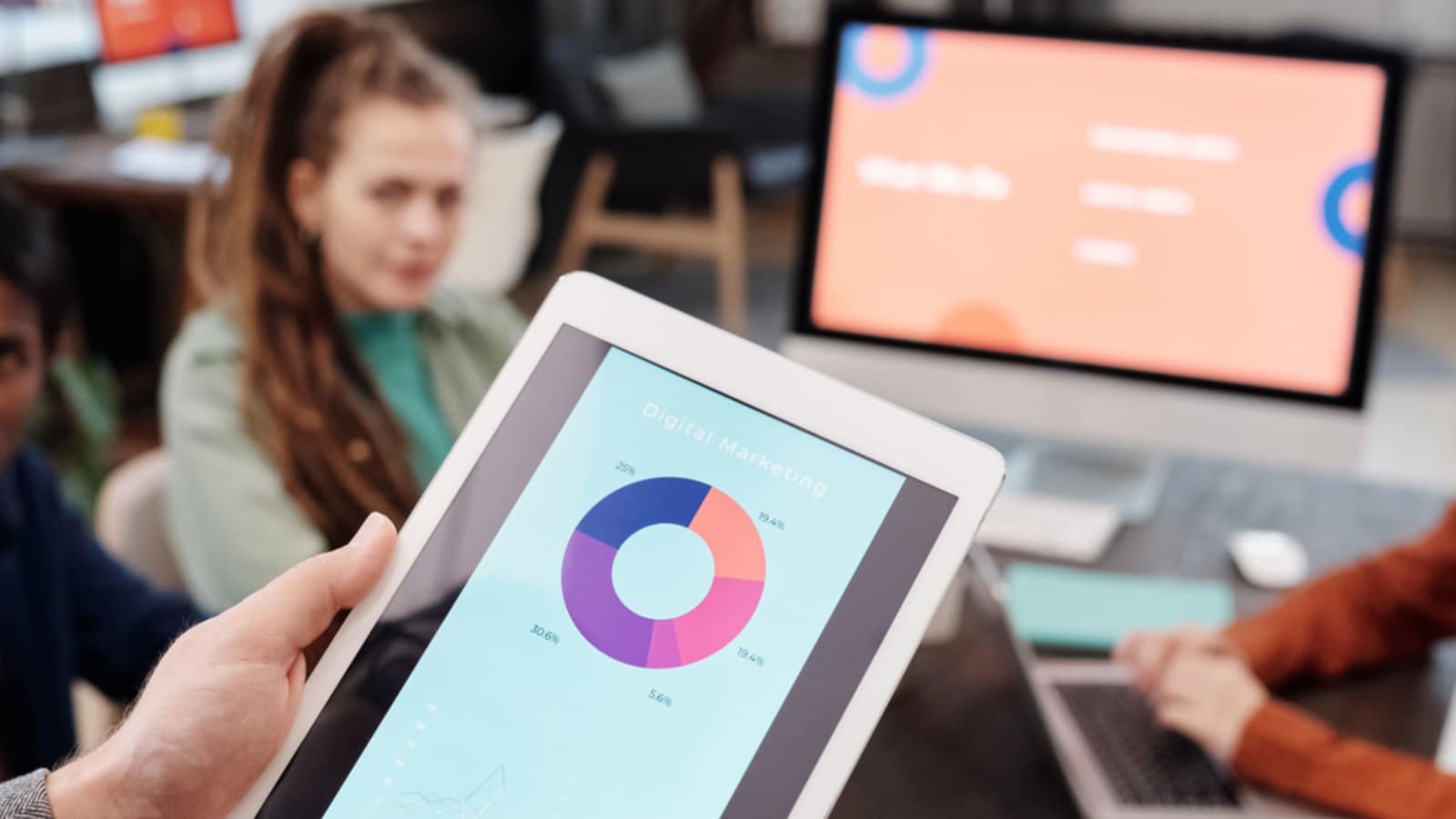 A graph on a tablet with a colleague and a screen  in the background