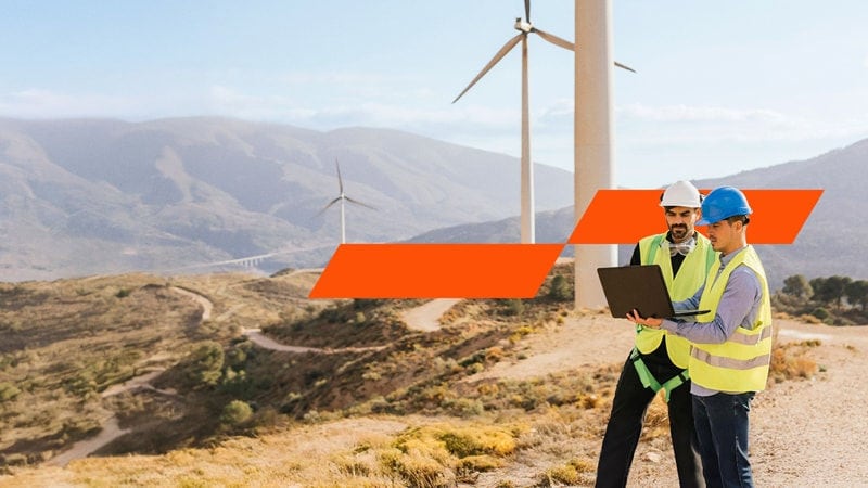 Two engineers in safety wear discussing over laptop in an open site with wind power generators in the background