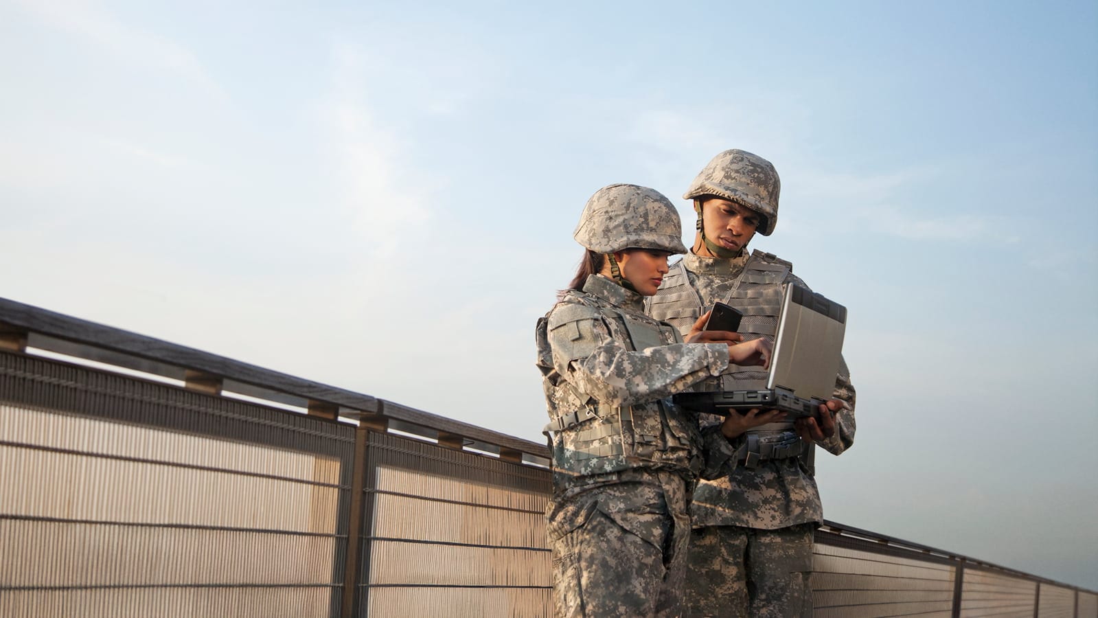 Two soldiers in camouflage uniforms and helmets stand outside next to a railing, with one holding a laptop and the other holding a smartphone while they both look at the laptop screen.