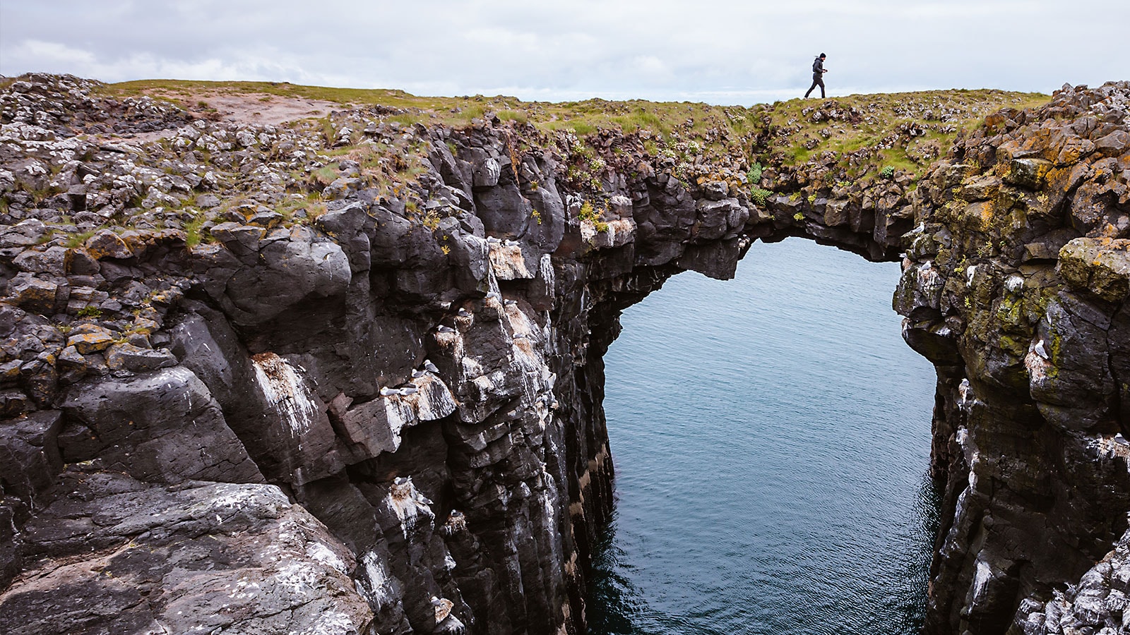 Man walking across a natural, stone bridge over water in Iceland