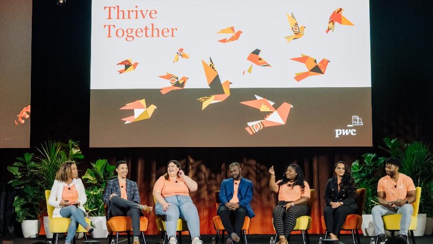 Image shows a group of 7 people from different ethnicities sitting on chairs on a stage for a panel discussion at a PwC Thrive Together event. There is a large screen behind them that says Thrive Together and has an image of birds flying in a flock.