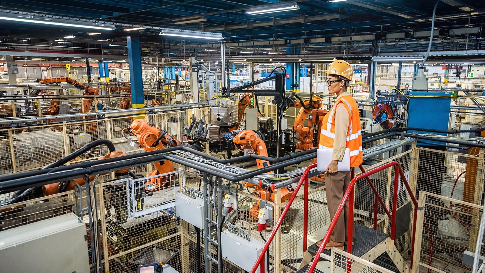 An engineer in a hard hat and safety vest oversees robotic arms on an automated industrial manufacturing floor, symbolizing the race to 2030 in smart factory innovation.