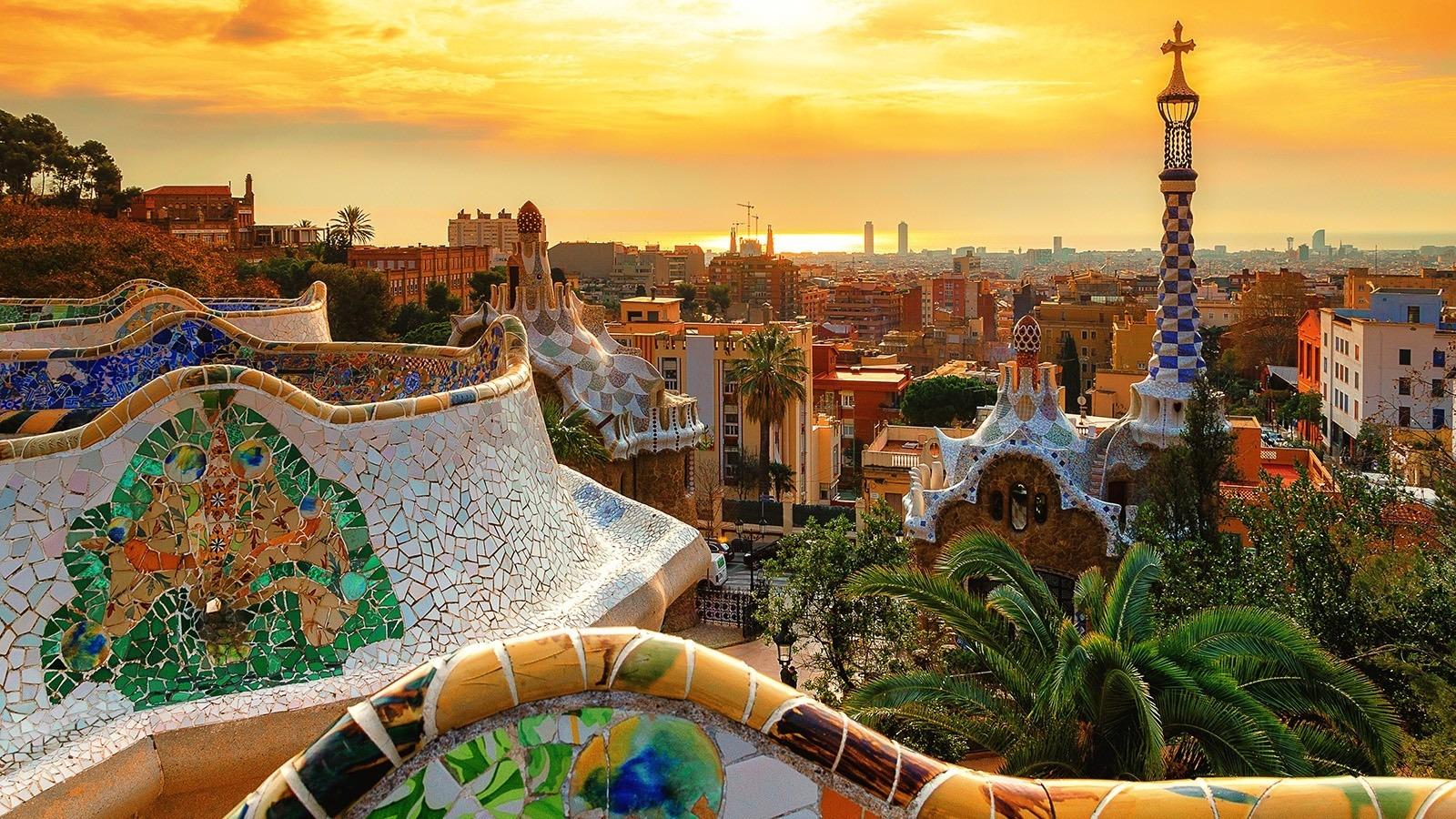 Colorful mosaic terraces of Park Güell in Barcelona at sunset, overlooking the city skyline and Mediterranean horizon.