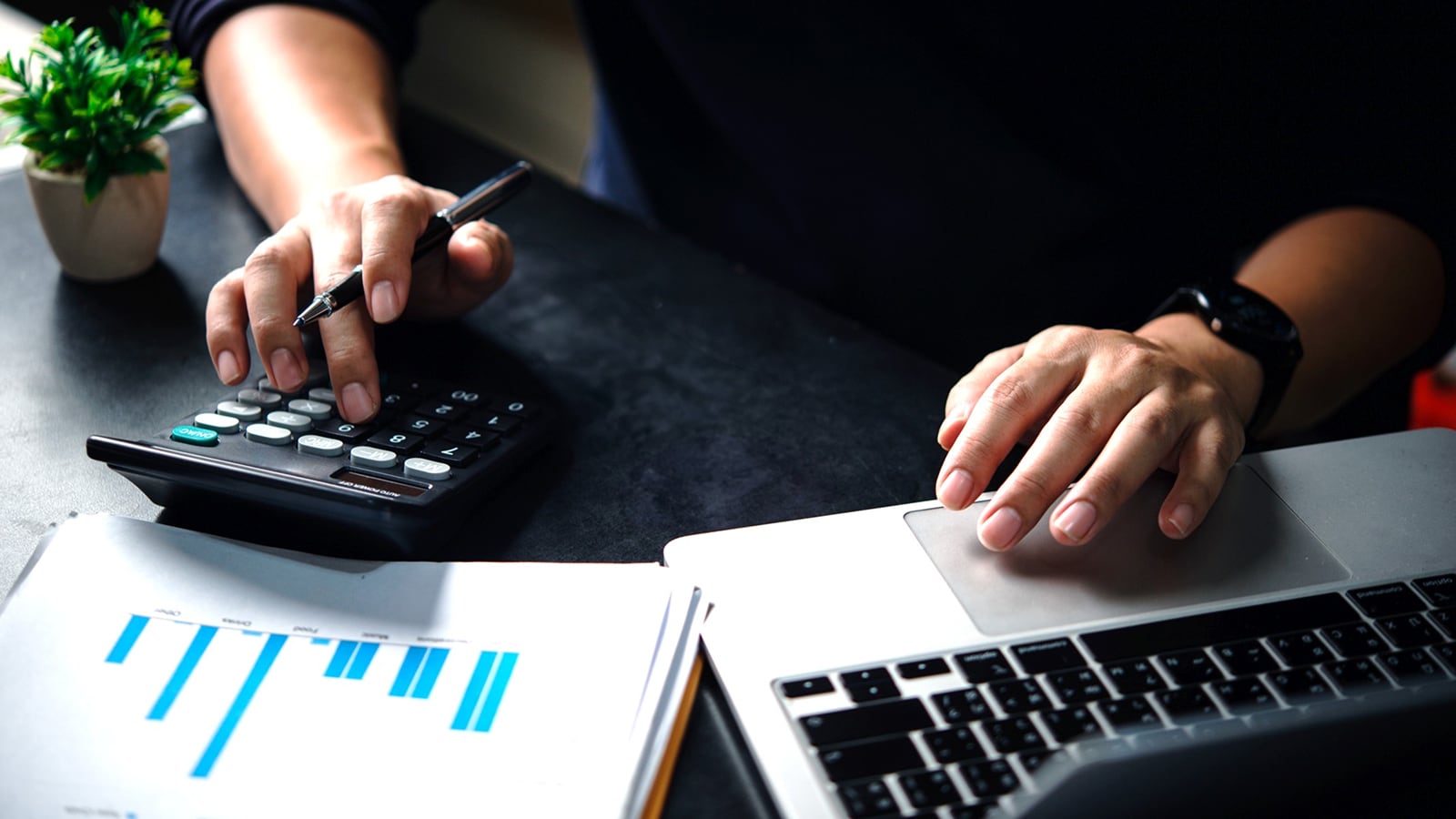 Hands of a person working on a desk. One hand on the computer and one on a calculator. 