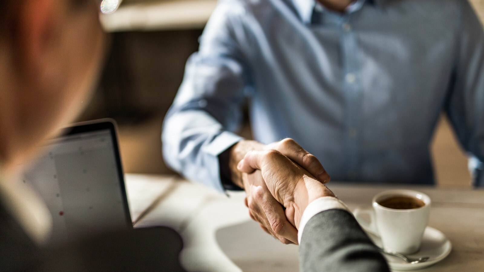 Two people shaking hand over a table. On the table there is a laptop and a cup of coffee