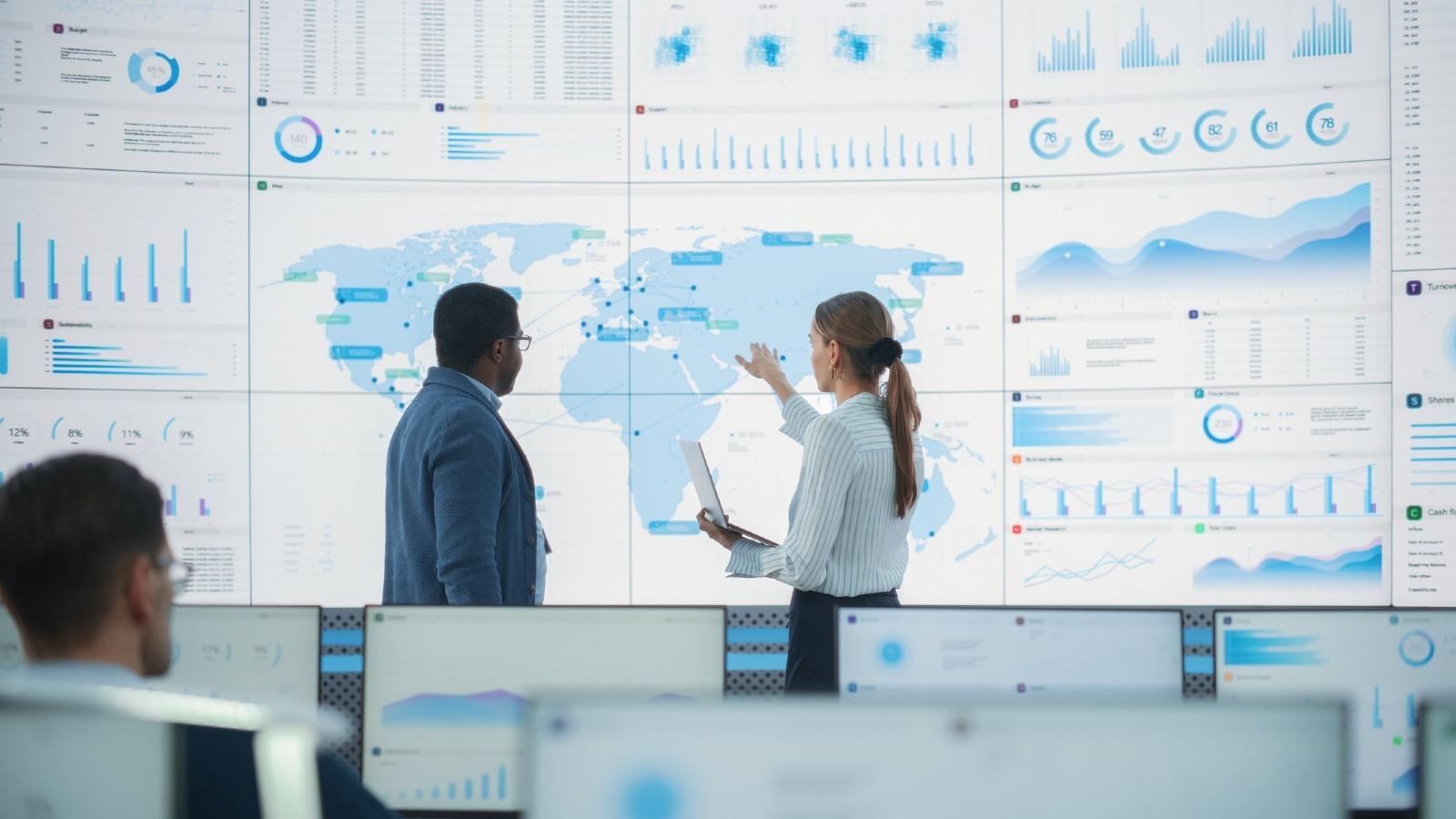 Two people standing in an office infront of a huge screen showing the world map